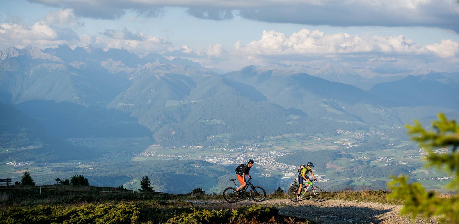 Zwei Mountainbiker fahren auf einem Bergpfad mit beeindruckender Alpenlandschaft im Hintergrund.