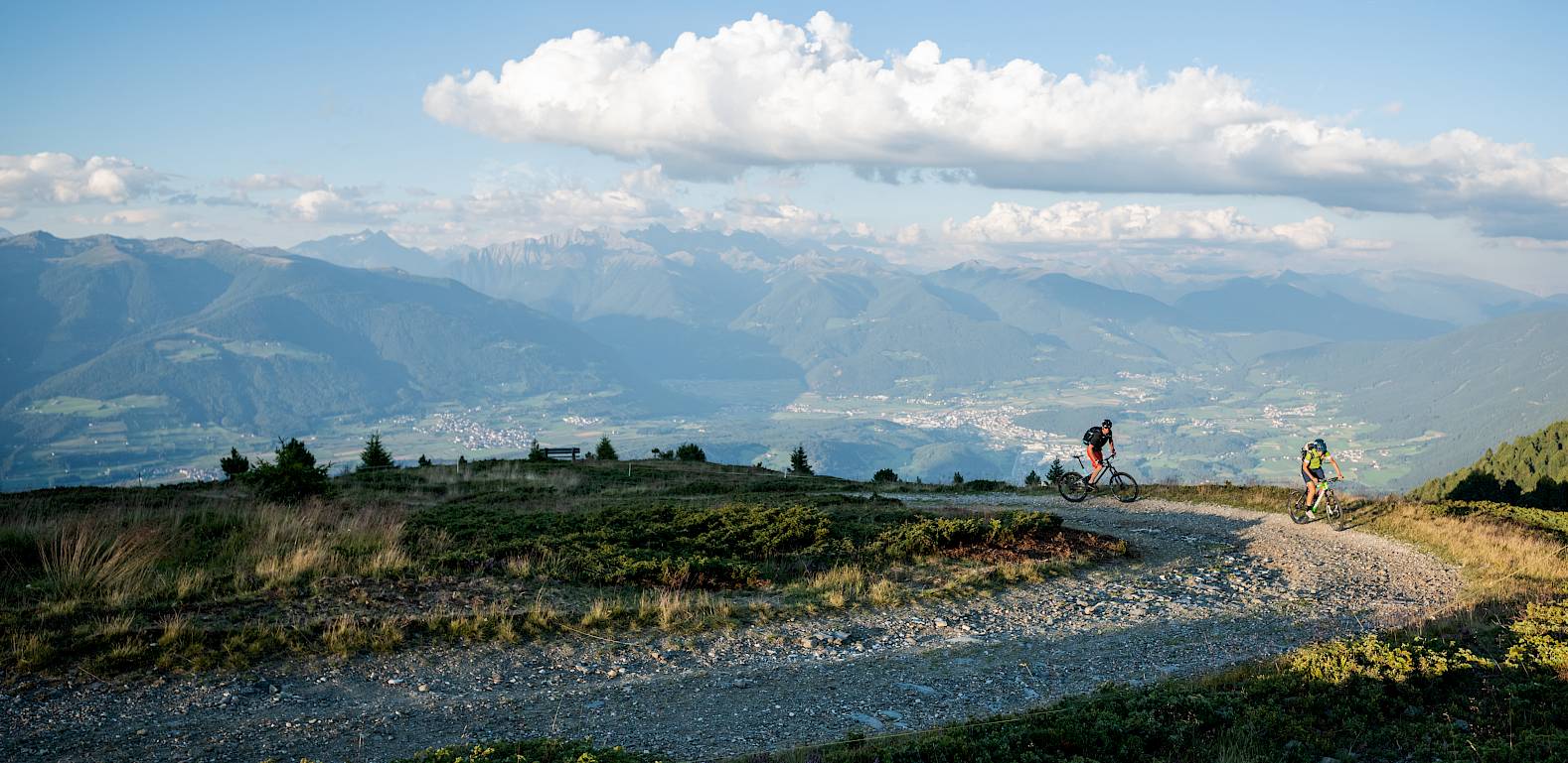 Zwei Mountainbiker fahren auf einem kurvigen Bergweg mit weitreichendem Alpenpanorama im Hintergrund.