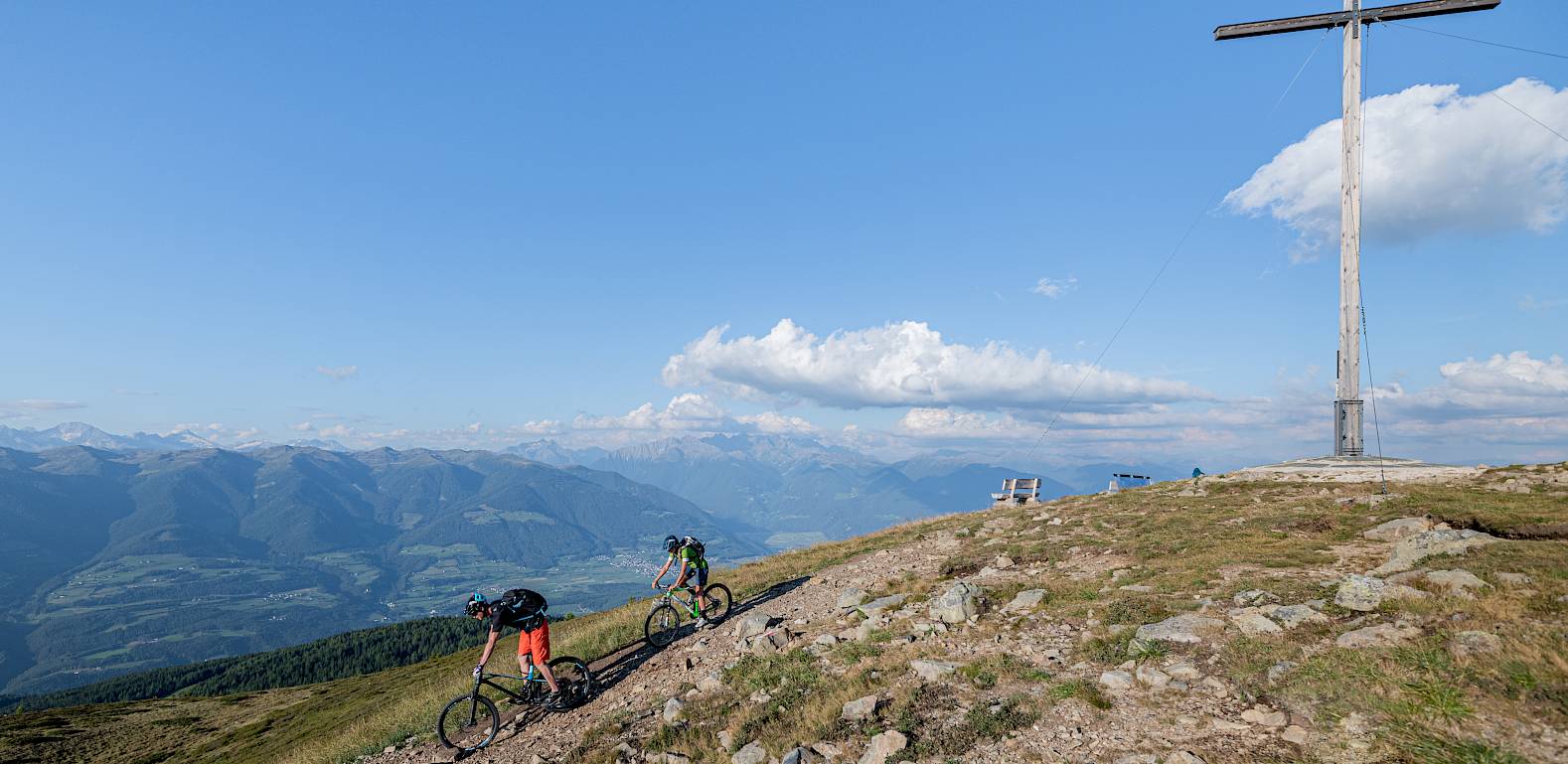 Zwei Radfahrer erklimmen einen Bergpfad neben einem großen Gipfelkreuz mit weitem Talblick.